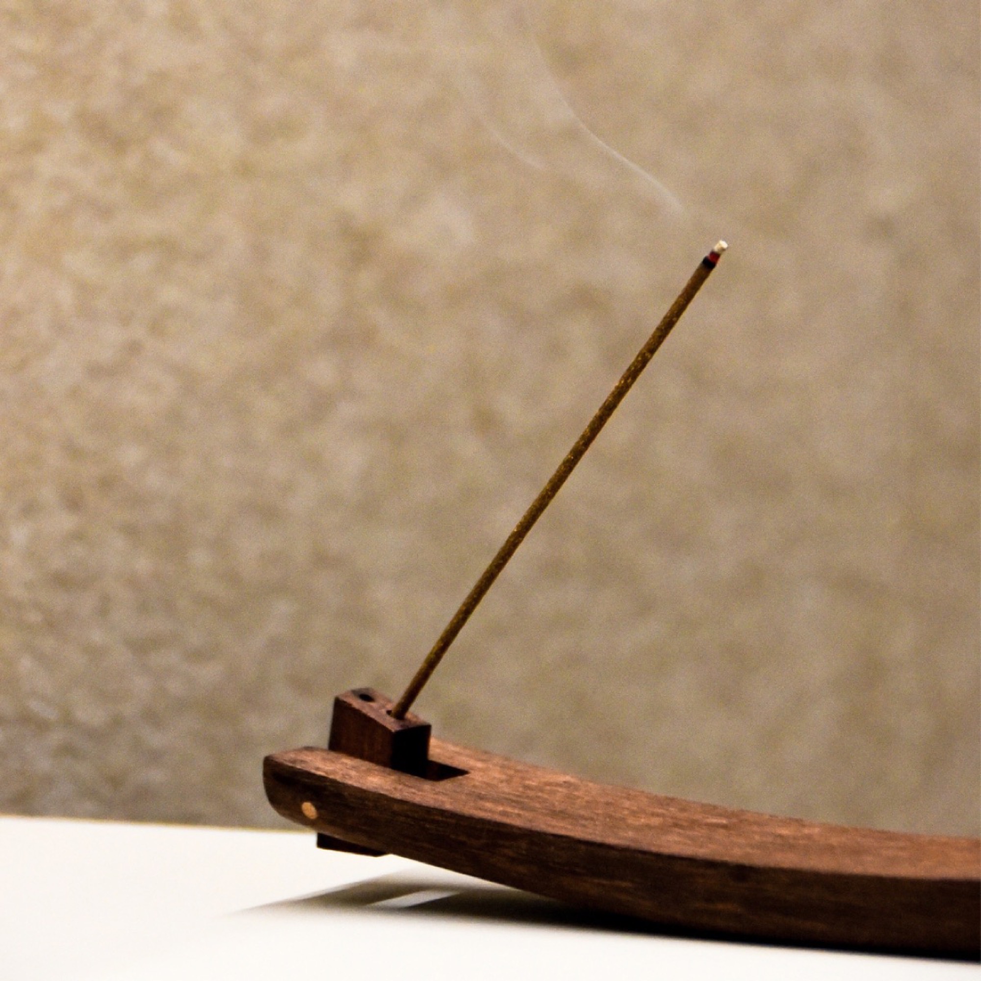 Close-up of the dark wooden incense holder, its stern joint pivoted out to hold a lit incense stick with smoke rising