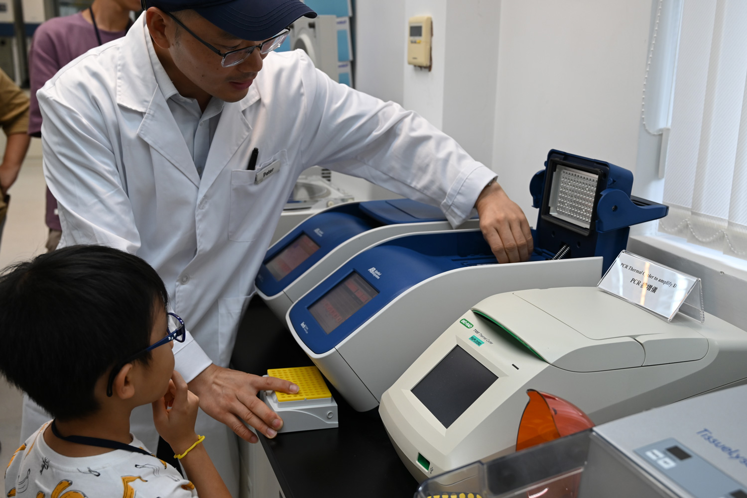 身穿白袍的本園科學人員在 PCR 擴增儀旁,向一名小朋友展示樣本盤