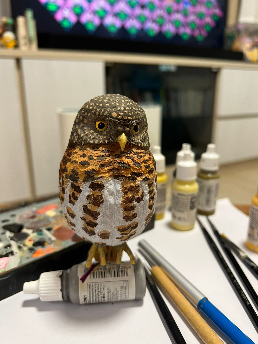 上色完成的斑頭鵂鶹模型,站在工作枱上,旁邊放着顏料瓶、畫筆和調色盤