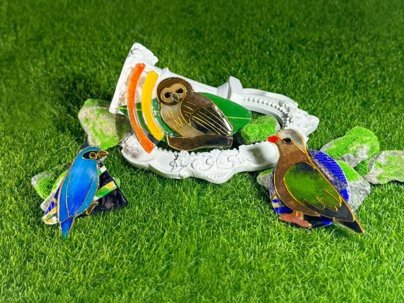 三件完成的雀鳥作品——貓頭鷹、藍色鶲鳥和綠藍色鳩鳥——以金線勾邊、填上通透色彩,放在仿草地上,旁邊有白色花紋相框和長了青苔的石頭